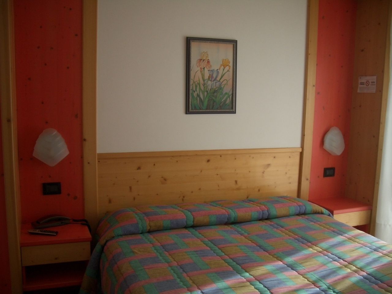 A bedroom with a wooden headboard, a colorful quilted bedspread, a bedside table with a lamp, and a framed floral artwork on the wall.