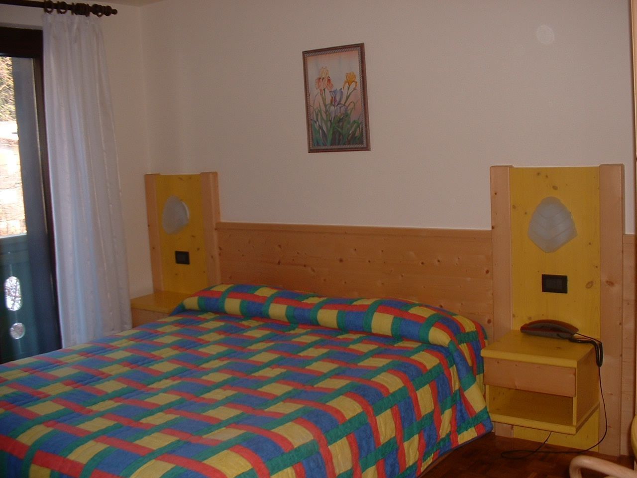 A bedroom with a wooden headboard, a colorful quilted bedspread, a bedside table with a telephone, and a wall-mounted picture.