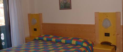 A bedroom with a wooden headboard, a colorful quilted bedspread, a bedside table with a telephone, and a wall-mounted picture.