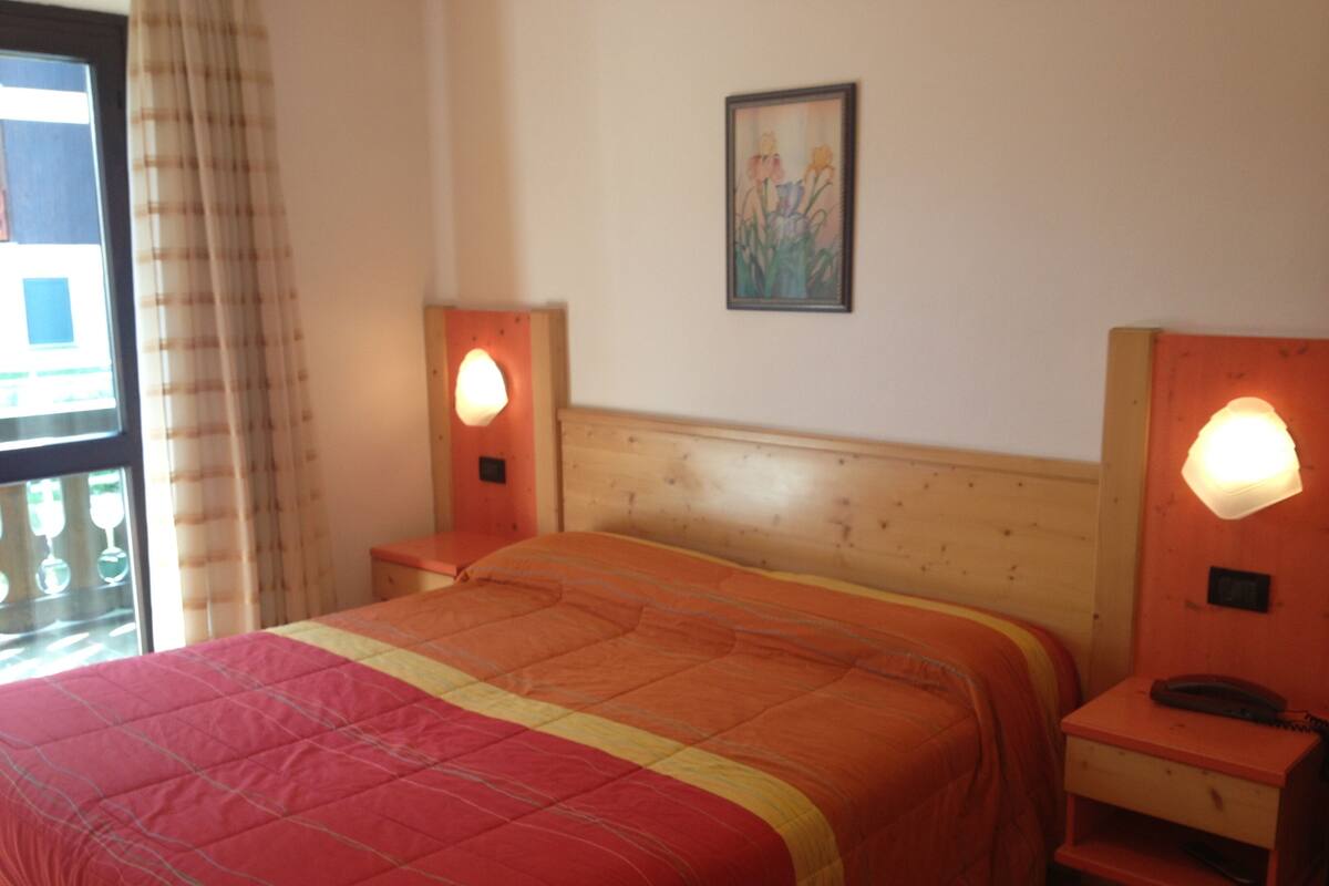 A hotel room with a wooden bed, two bedside tables, a nightstand, a window with curtains, and a wall-mounted picture.