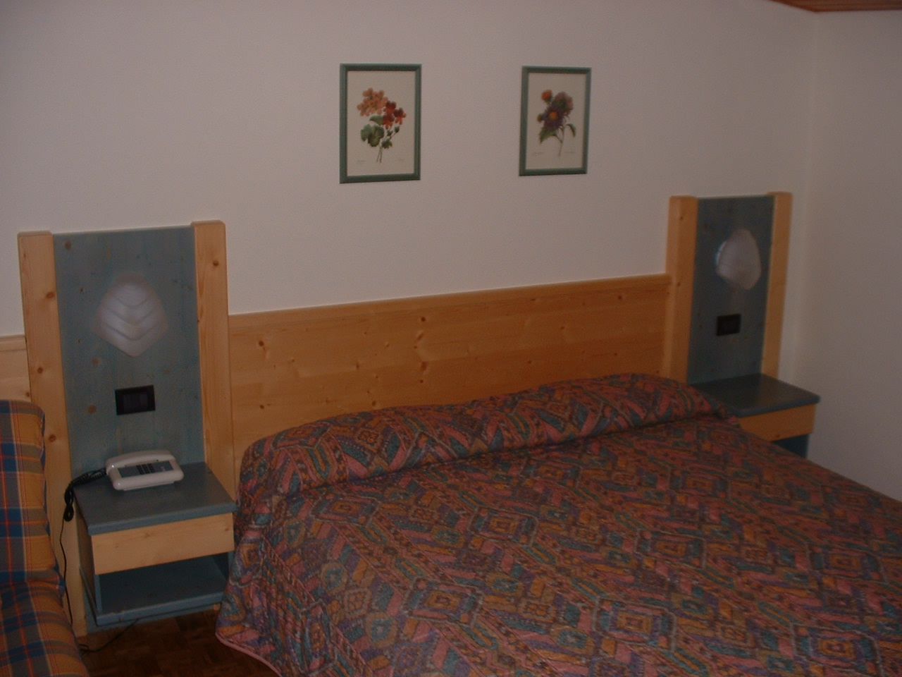 A bedroom with a wooden headboard, a patterned bedspread, and two bedside tables with a small lamp and a telephone on one.
