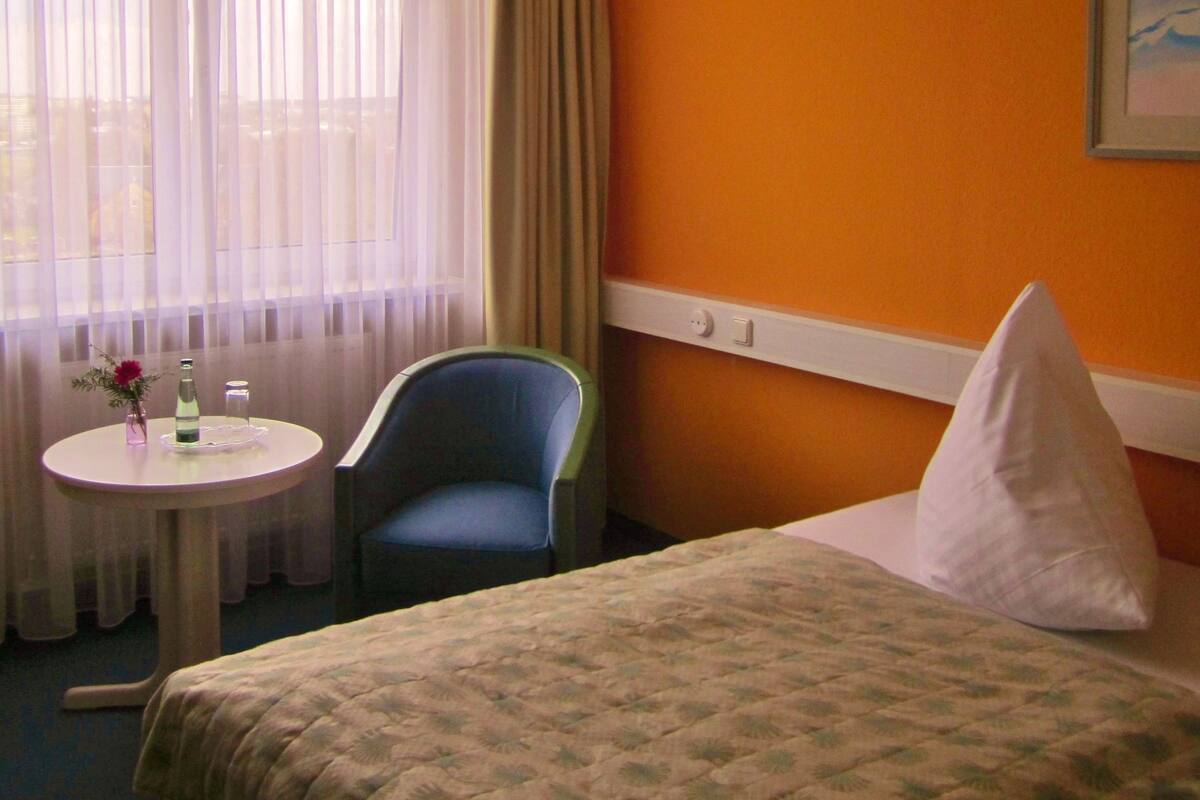 A hotel room with a bed, a chair, a small table with a vase and a bottle, and a window with curtains.