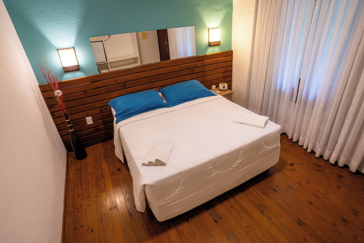 A neatly made bed with a blue pillow and white linens, a wooden headboard, a nightstand with a lamp, and a window with white curtains.