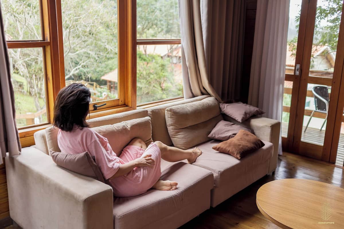 A person sitting on a sofa in a room with large windows, a wooden floor, and a round table.