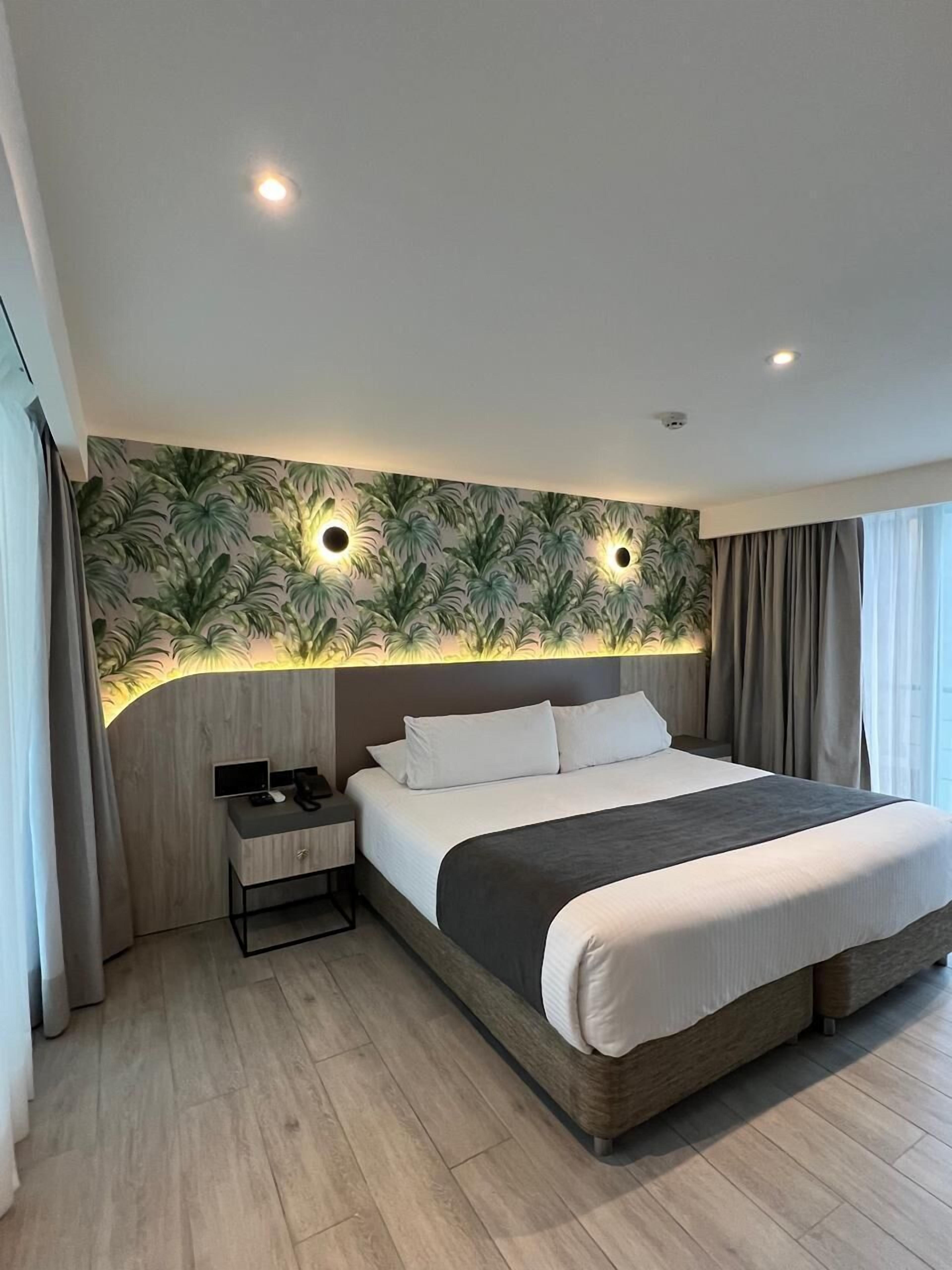 Una habitación de hotel moderna con una cama grande, una mesita de noche con un teléfono y una tableta, una pared con estampado de hojas tropicales e iluminación empotrada en el techo.