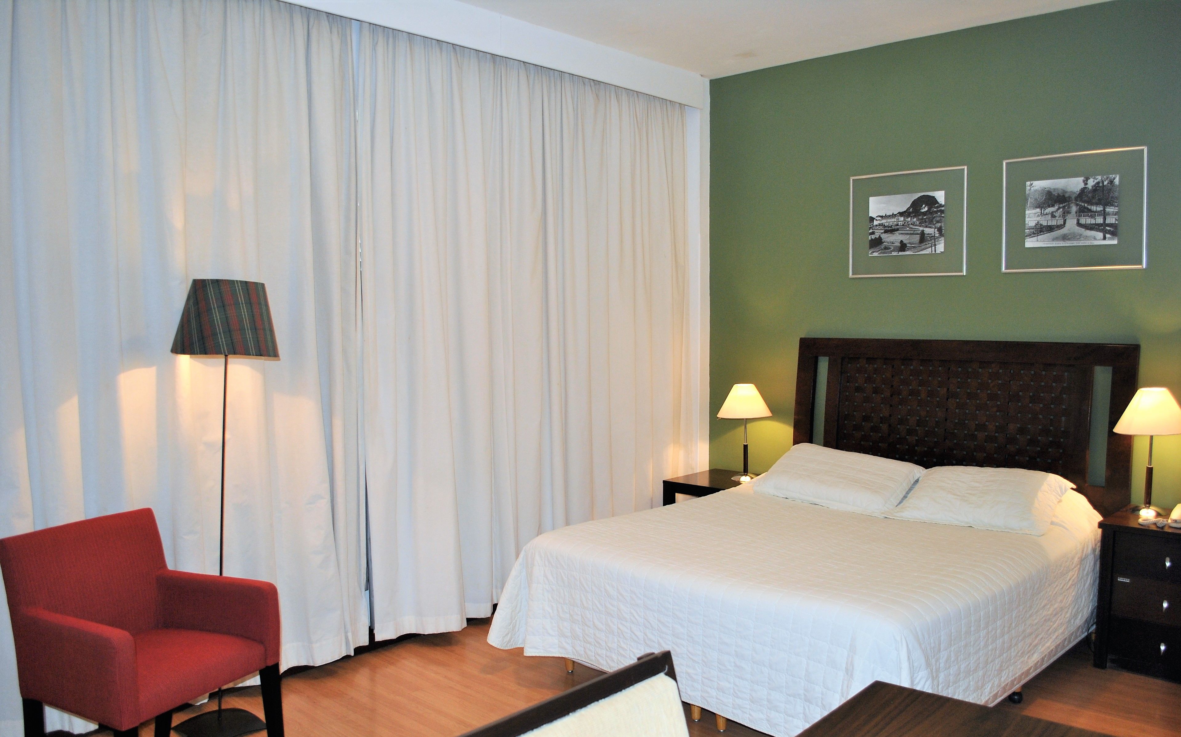 A hotel room with a bed, two bedside tables with lamps, a red armchair, and a large window with white curtains.