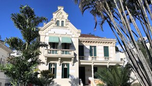 Un edificio de dos pisos con balcón, una escalera y un jardín con palmeras.