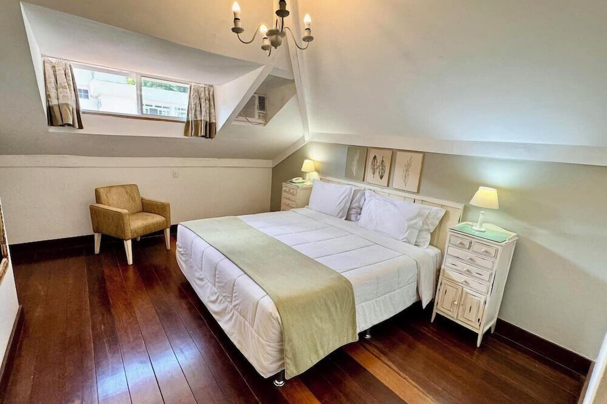 Ein Schlafzimmer mit einem Bett, einem Sessel, einem Nachttisch, einer Lampe, einem Kronleuchter und einem Fenster mit Vorhängen.