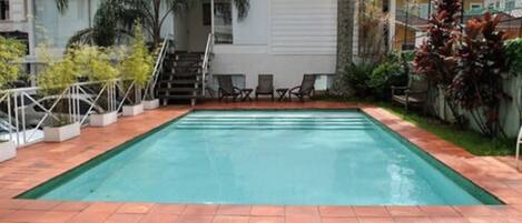 A rectangular swimming pool with a tiled deck, surrounded by a fence and various plants.