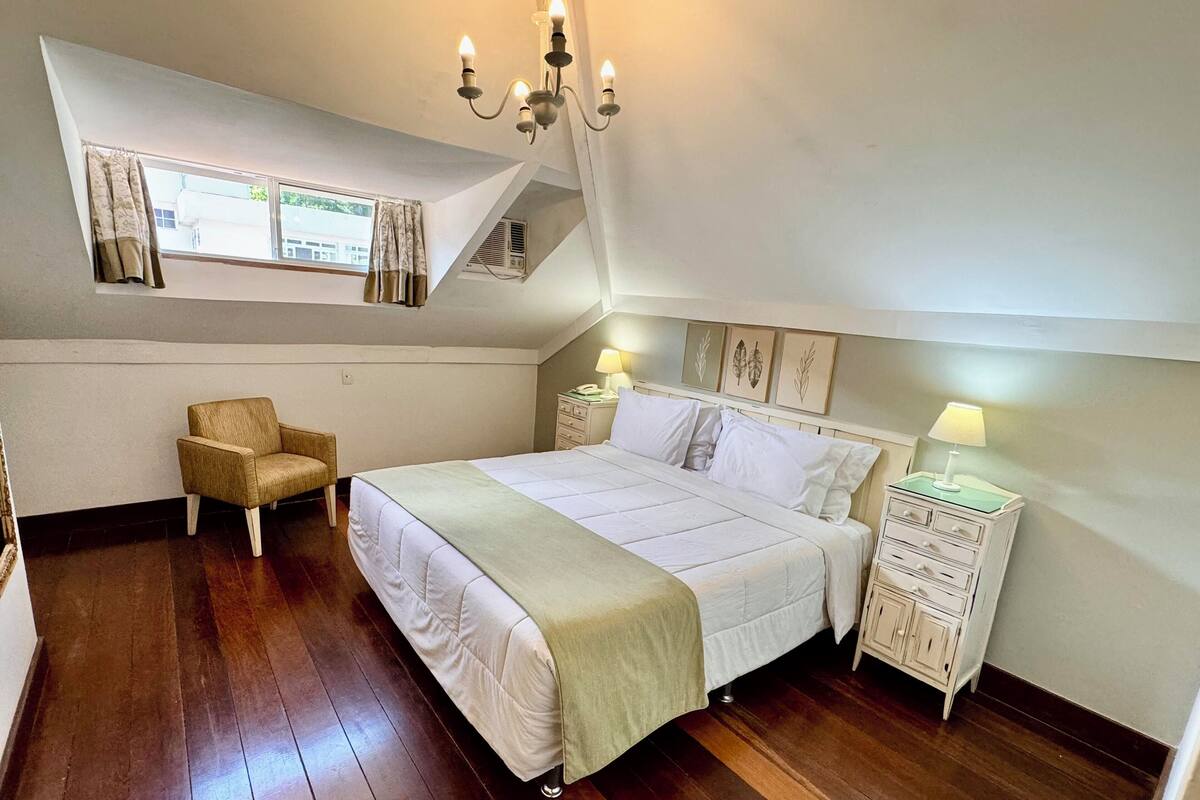 Ein Schlafzimmer mit Bett, Stuhl, Nachttisch, Lampe und einem Fenster mit Vorhängen.