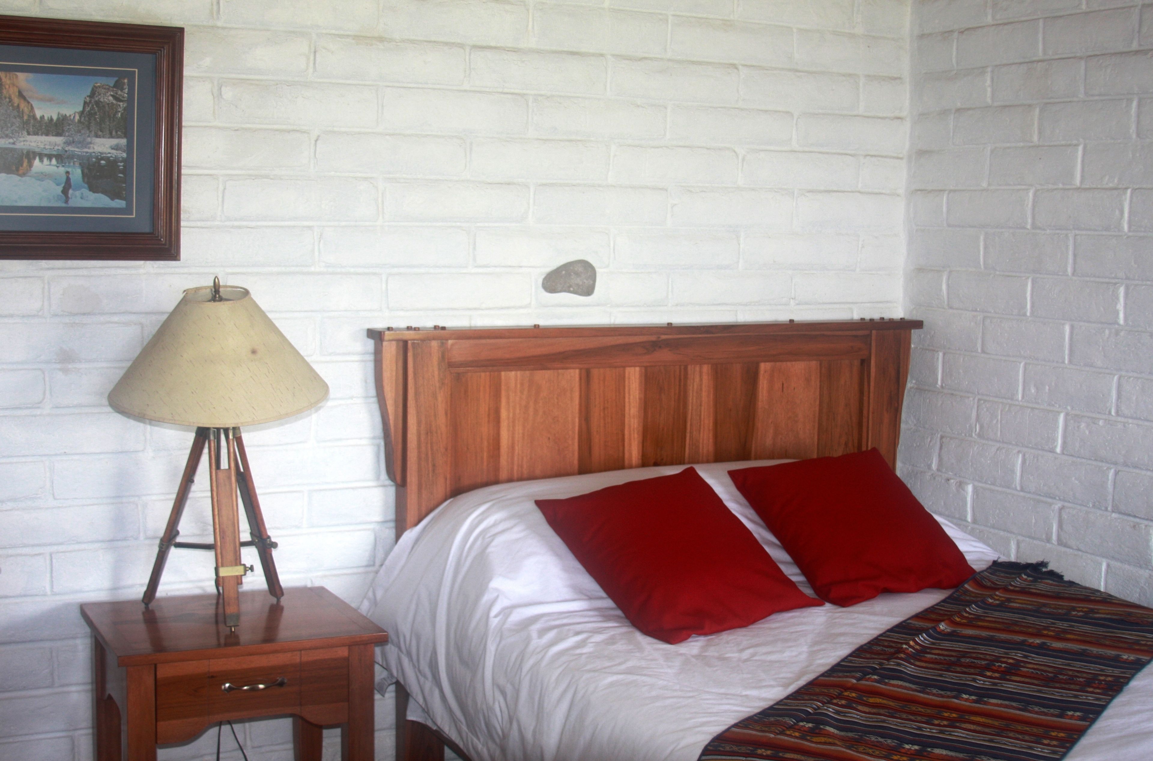 A bed with white bedding and red pillows, a wooden bedside table with a lamp, and a framed picture on the wall.