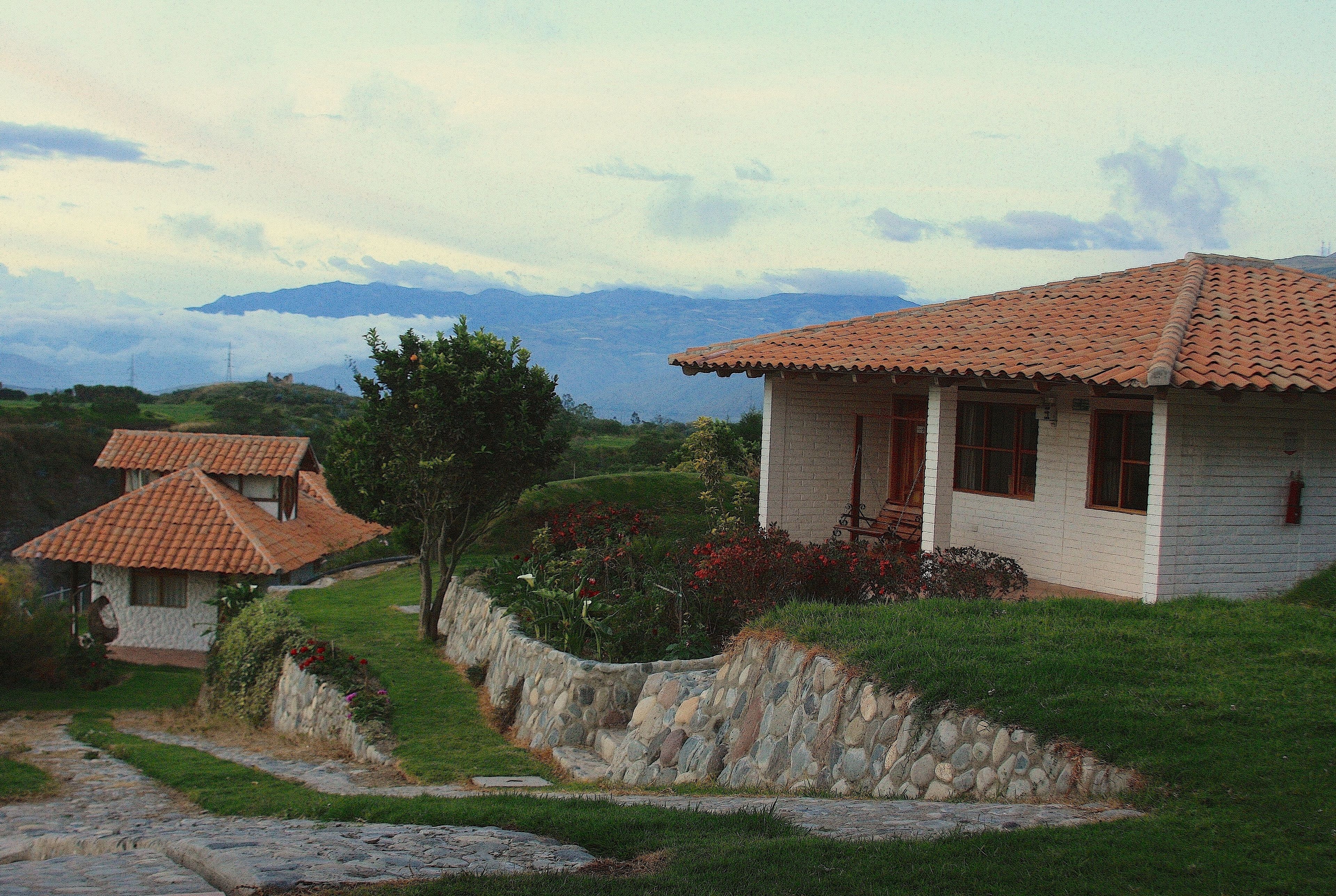 Una casa de dos pisos con un muro de piedra y techo de tejas, rodeada por un jardín y un camino de piedra.