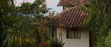A rustic house with a tiled roof, surrounded by lush greenery and a clear sky.