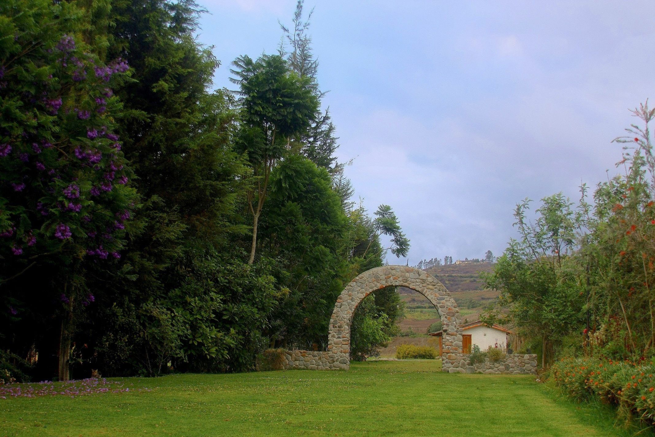 Un arco de piedra en un jardín exuberante, con vistas a una colina y un edificio pequeño al fondo.