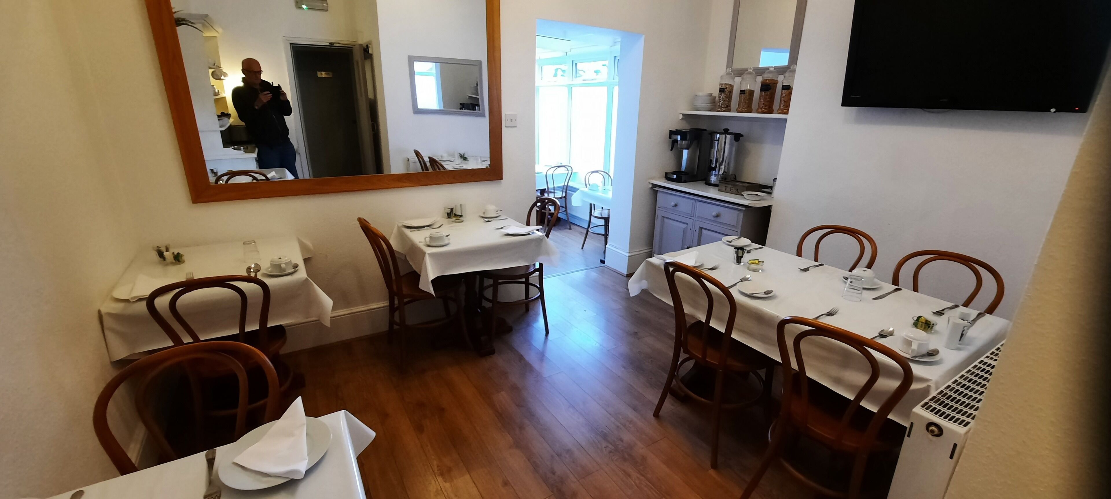 A dining area with tables set for four, a coffee machine, and a television on the wall.