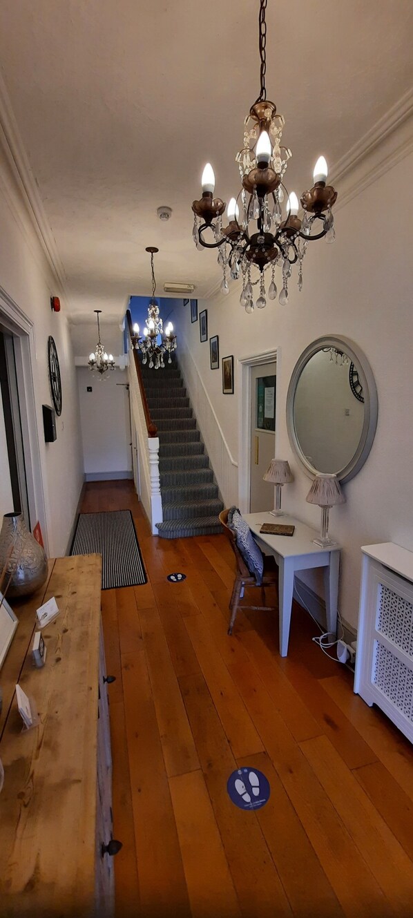 A hallway with wooden flooring, a staircase, a chandelier, a mirror, and a small table with a lamp. - Colebrook Guest House (Farnborough)