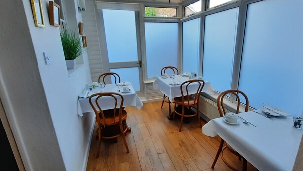 A dining area with four tables, each set with white tablecloths and wooden chairs. The room has a wooden floor and large windows with blue-tinted glass. - Colebrook Guest House (Farnborough)