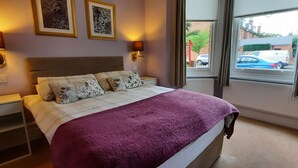 A bedroom with a bed, two wall-mounted lamps, a window with a view of parked cars, and a door. - Colebrook Guest House (Farnborough)