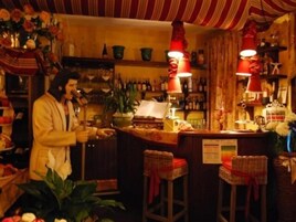 Un bar con estantes de madera llenos de botellas, una barra con caja registradora y taburetes de mimbre.
