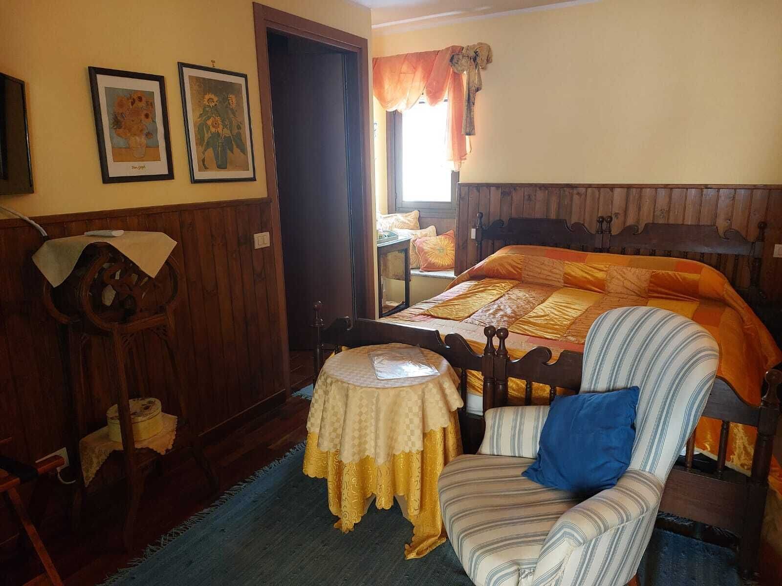 A bedroom with a wooden bed, a chair, a small table, and two framed pictures on the wall.