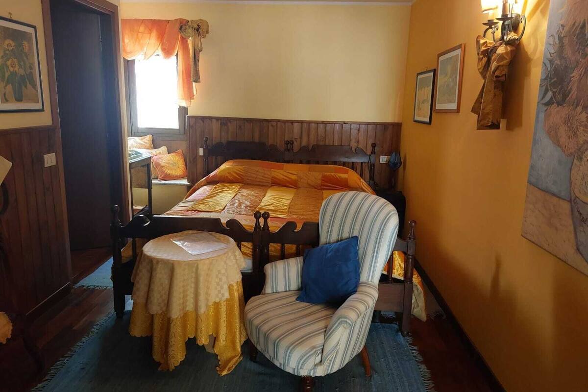A bedroom with a wooden bed, a striped armchair, a small round table, and a window with curtains.