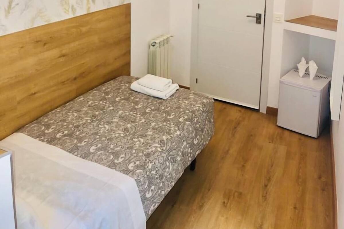 A bedroom with a wooden headboard, a patterned bedspread, a white bedside table with a tissue box, a radiator, a white door, and a mounted TV.