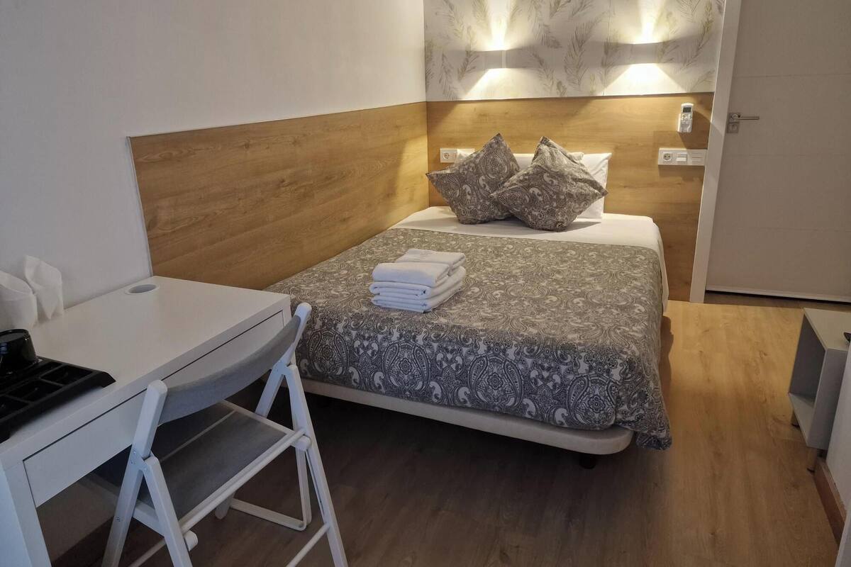A compact hotel room with a bed, bedside table, and a desk with a chair.