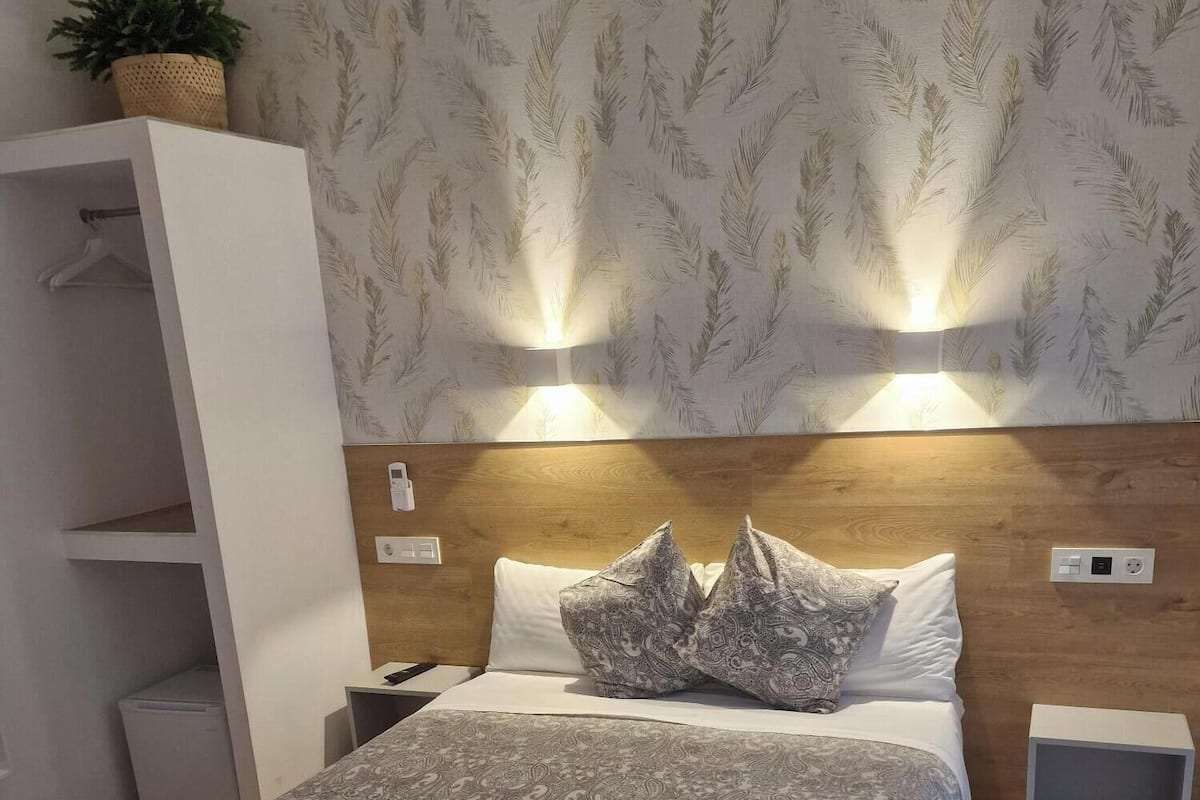 A neatly made bed with patterned bedding, a bedside table with a lamp, and a wall-mounted shelf with a plant.