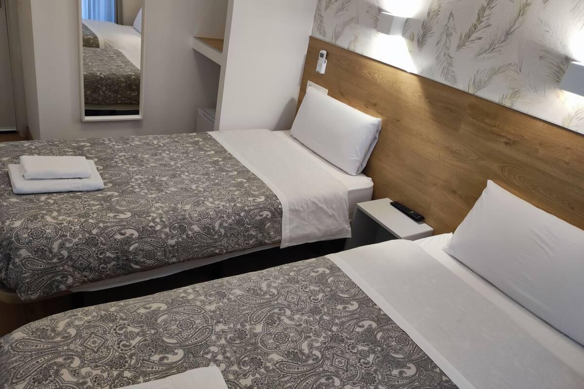 A hotel room with two beds, each with a patterned bedspread and white pillows. There is a bedside table with a remote control and a wall-mounted light fixture.