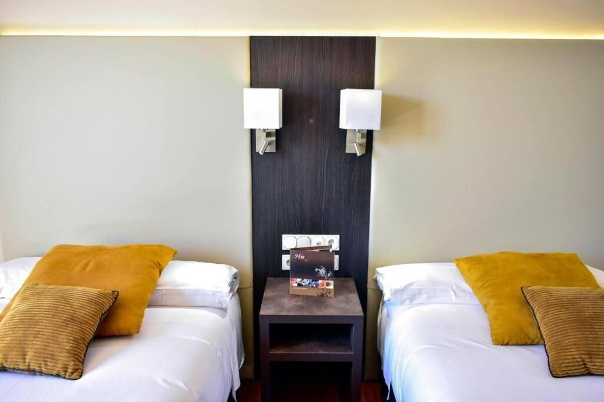 A hotel room with two beds, each with a white mattress and a mustard-colored pillow. There is a small wooden nightstand between the beds with a white lamp and a small box on it.
