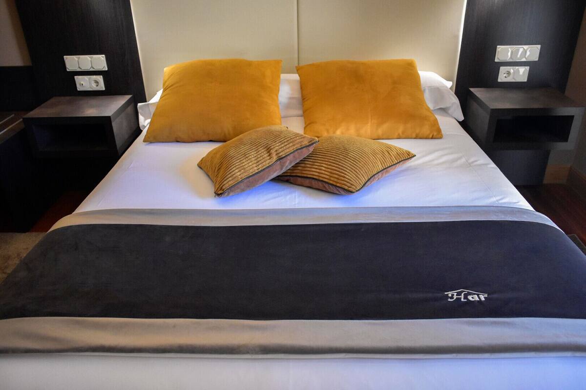 A neatly made bed with two large pillows and a folded blanket, a bedside table with a lamp, and a wall-mounted light switch.