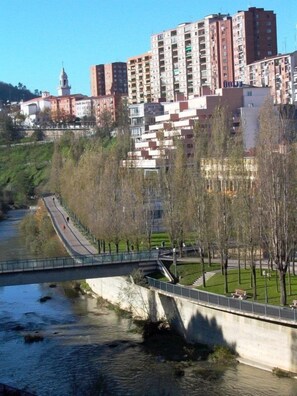 A riverside walkway with a bridge, surrounded by residential buildings and a park with trees.
