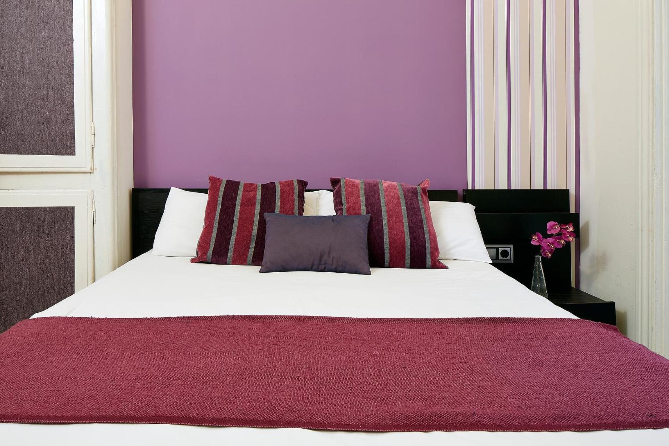 Cama bem arrumada com manta vermelha, almofadas brancas e almofadas listradas em quarto com paredes roxas e cortina listrada.