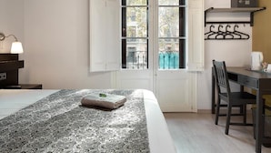 A hotel room with a bed, a desk, a chair, a hanging clothes rack, and a window with a view of buildings outside. - Brustar Gòtic (Barcelona)