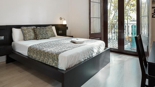 A modern bedroom with a large bed, a nightstand with a lamp, and a view of the outdoors through a glass door. - Brustar Gòtic (Barcelona)