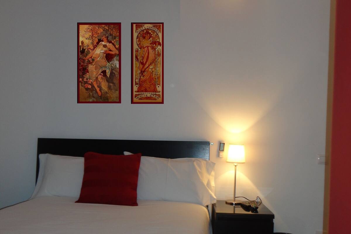 A hotel room with a bed, a nightstand with a lamp, and two wall-mounted artworks.