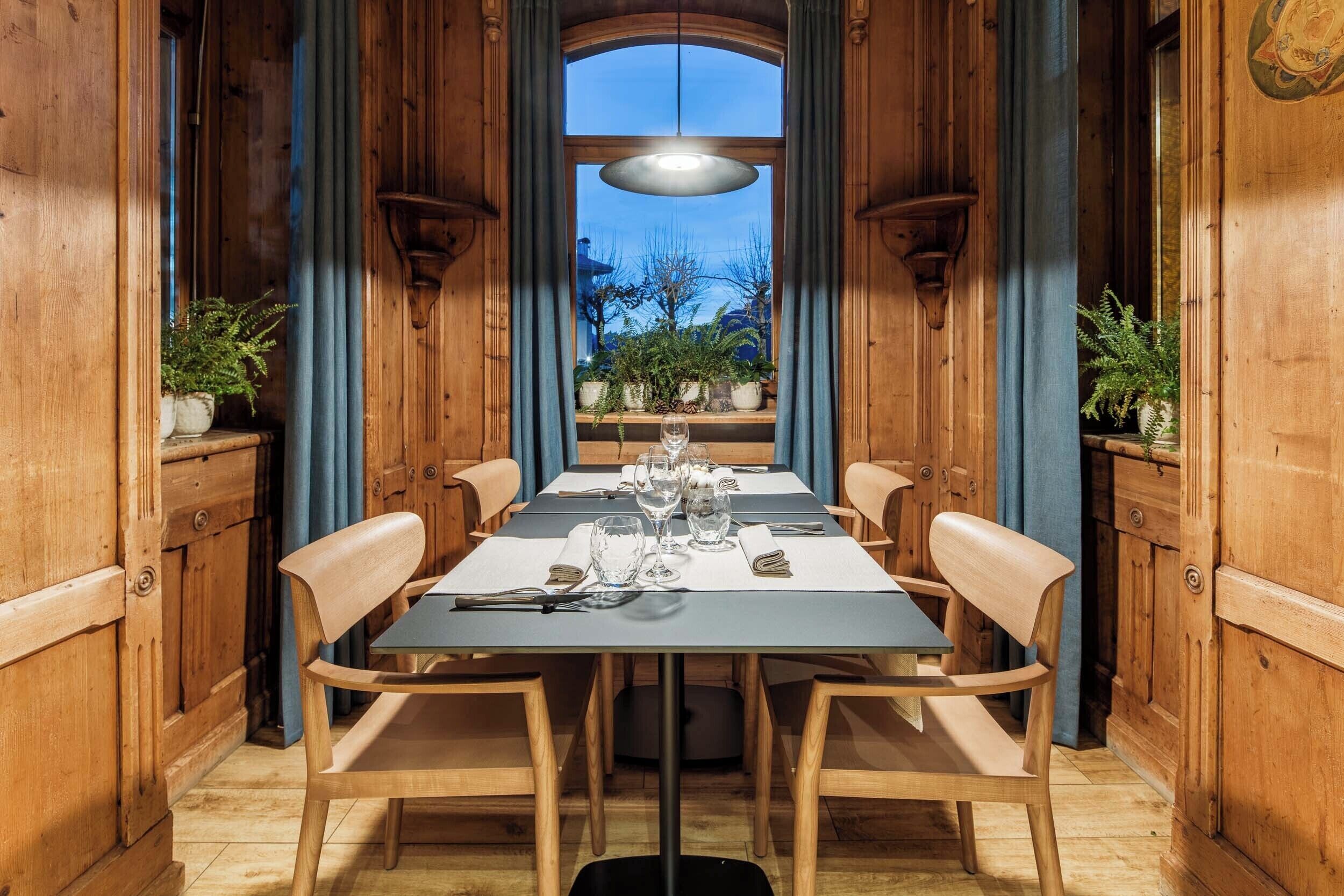 A dining area with a long table set for four, flanked by wooden chairs, with a view of the outdoors through a large window.