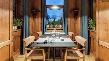A dining area with a long table set for four, flanked by wooden chairs, with a view of the outdoors through a large window.