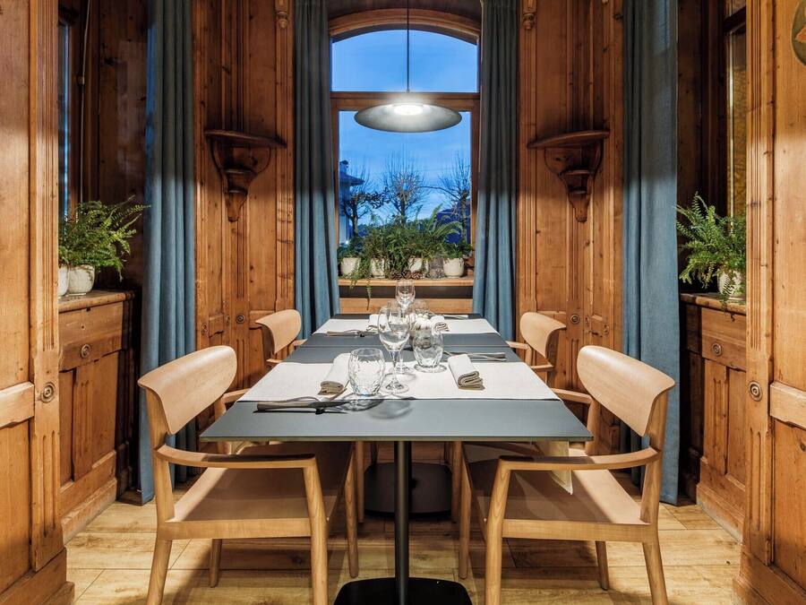 A dining area with a long table set for four, flanked by wooden chairs, with a view of the outdoors through a large window.