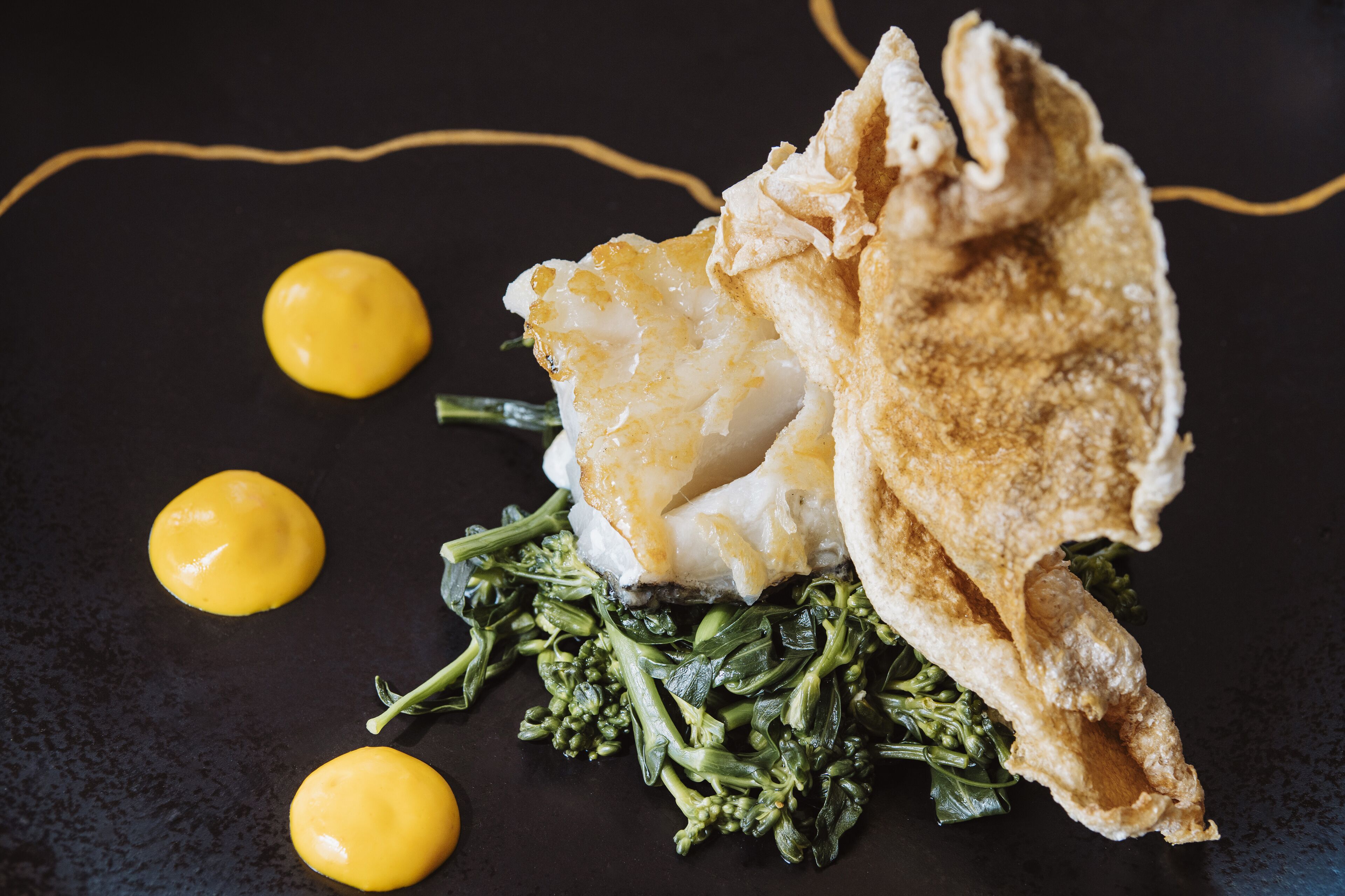 A dish with fish, greens, and a crispy element on a dark plate.