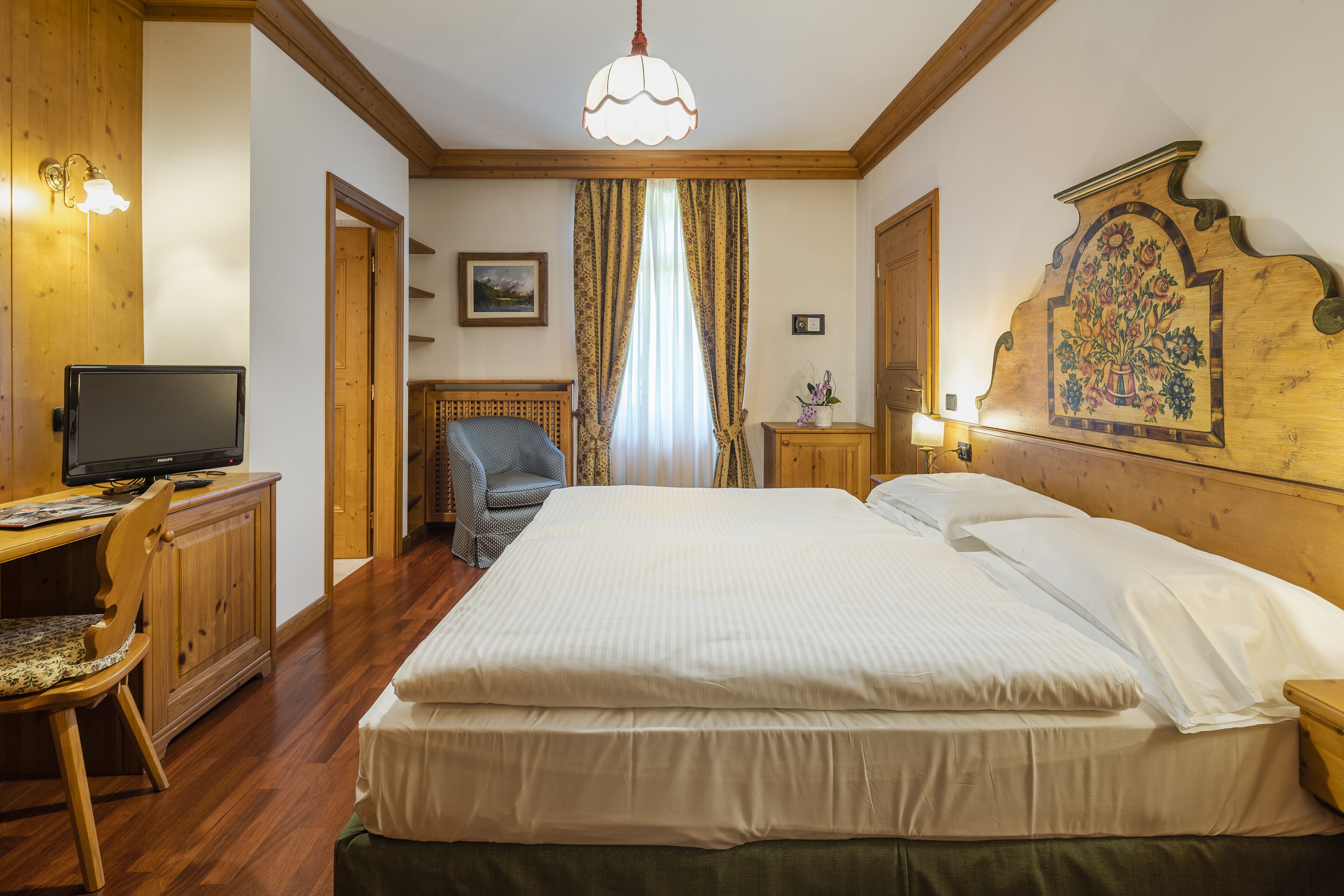 A hotel room with a bed, a desk with a computer, a chair, and a wooden headboard with a floral design.