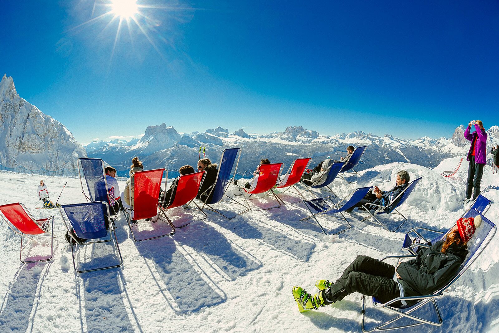 A snowy mountain scene with several people sitting on sun loungers, overlooking a range of mountains under a clear sky.