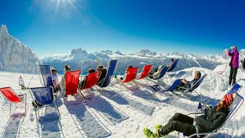 Un paysage de montagne enneigé où l’on voit plusieurs personnes assises sur des chaises longues, contemplant une chaîne de montagnes sous un ciel dégagé.