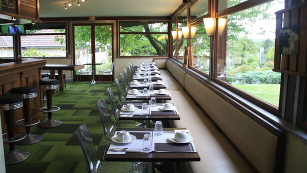 Een restaurant met lange tafels klaar voor diners, grote ramen met uitzicht op groen en een bar met krukjes.