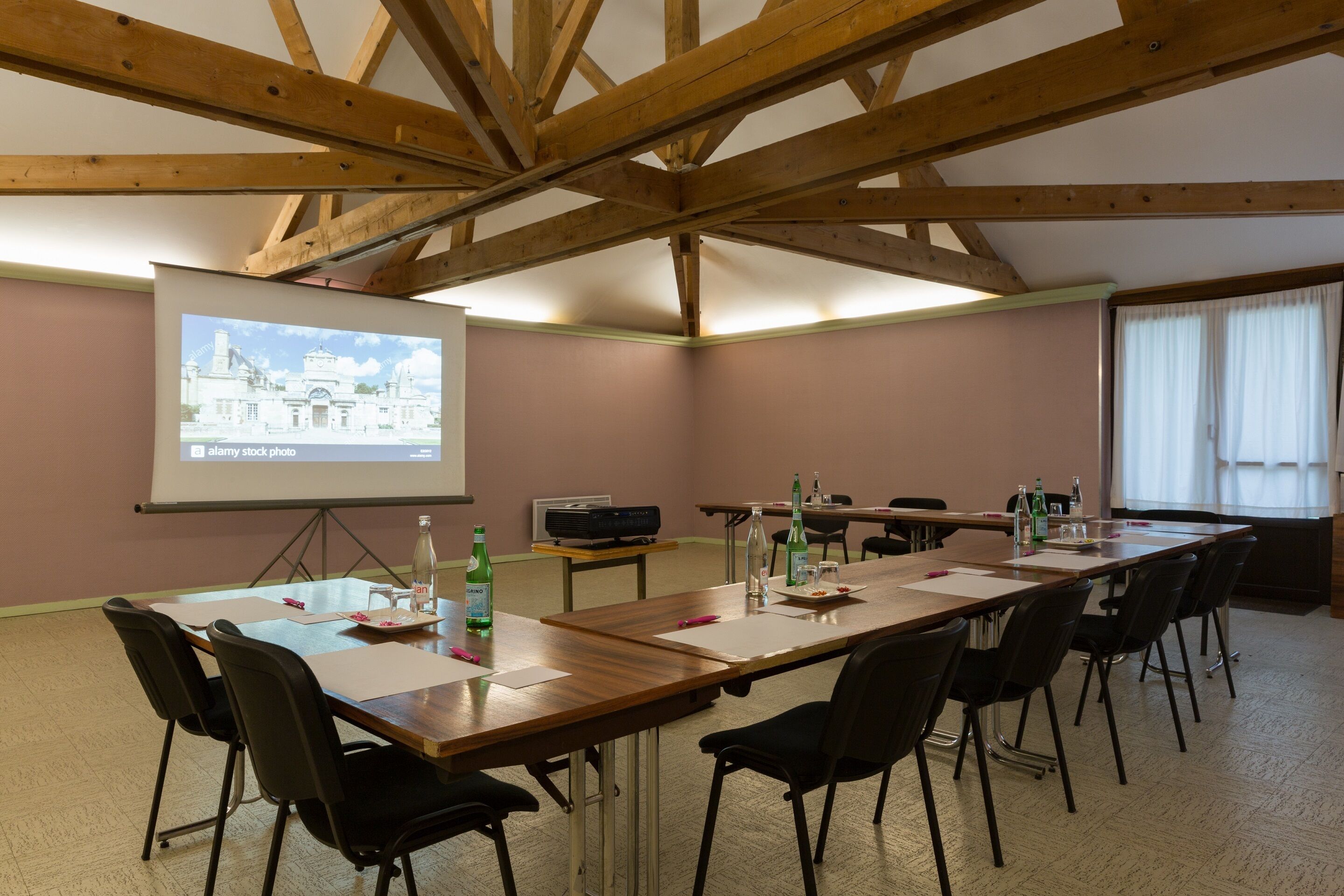 Un salón de conferencias con una larga mesa, sillas, una pantalla de proyector y un ventanal con cortinas.