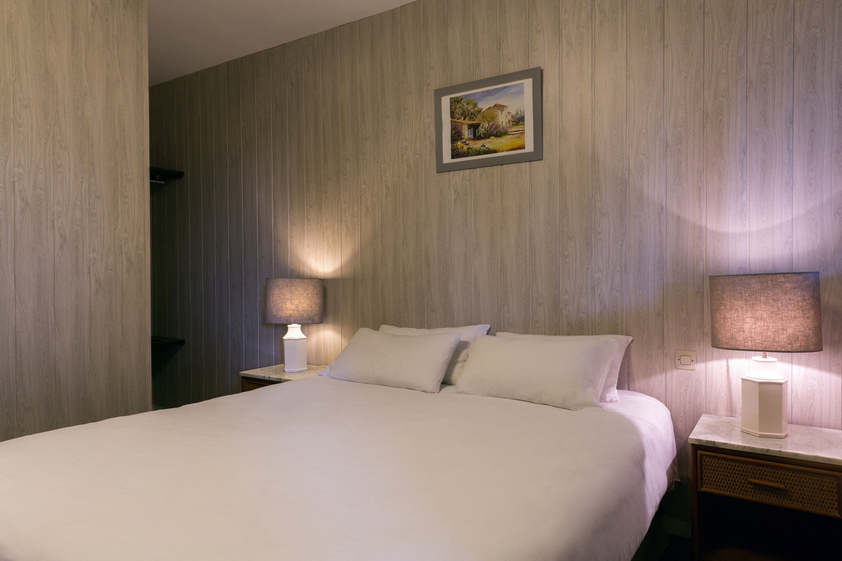 A neatly made bed with white linens, two bedside lamps, a framed picture on the wall, and a wooden headboard.