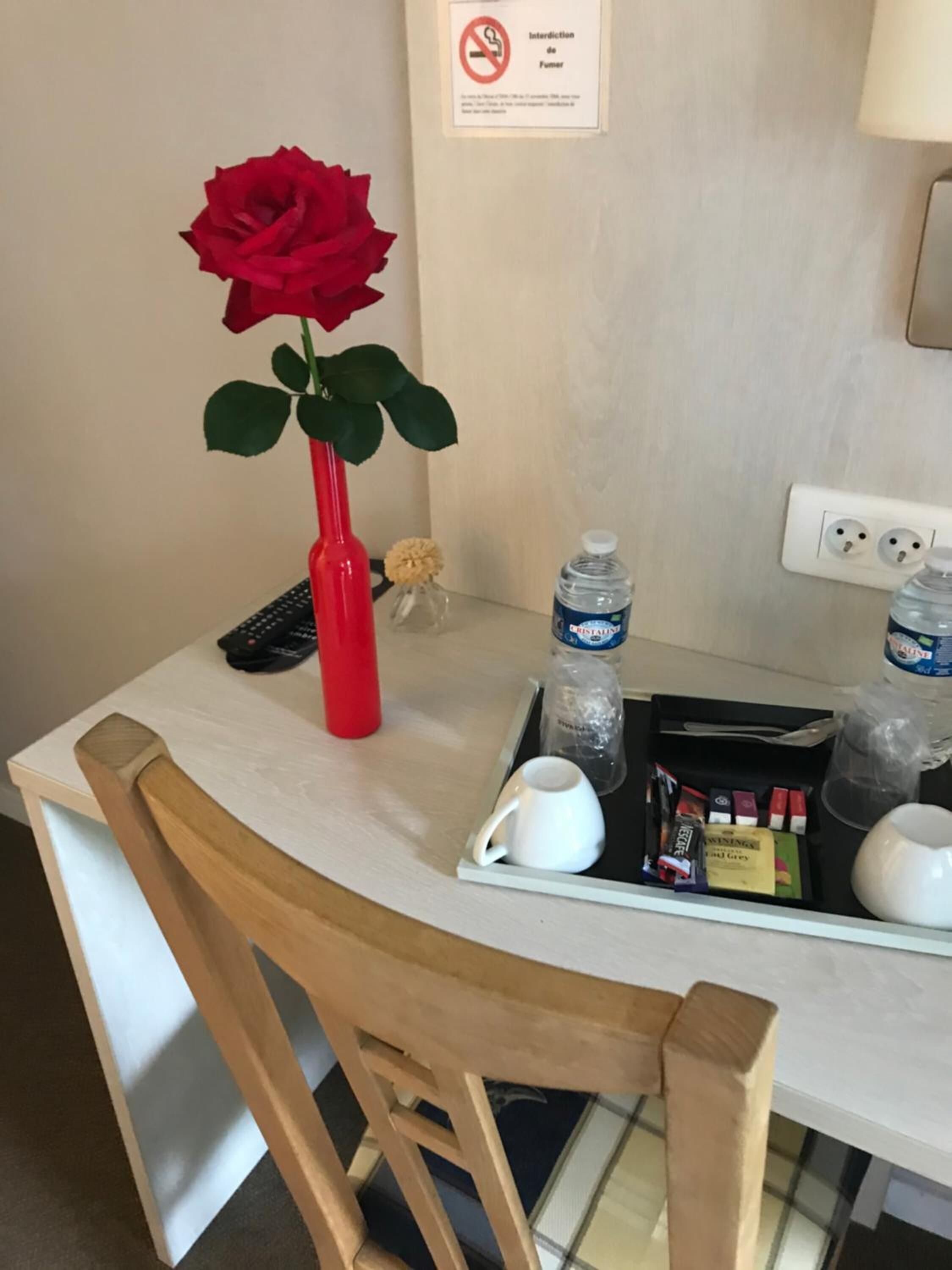 A hotel room with a wooden chair, a white table, a red vase with a rose, a small potted plant, a water bottle, a glass of water, a white mug, a tray with snacks, and a sign prohibiting smoking.