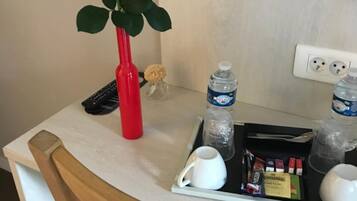 A hotel room with a wooden chair, a white table, a red vase with a rose, a small potted plant, a water bottle, a glass of water, a white mug, a tray with snacks, and a sign prohibiting smoking.