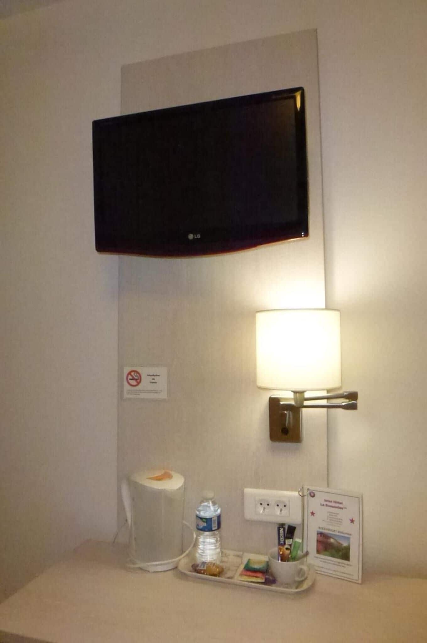 A hotel room with a flat-screen TV mounted on the wall, a bedside lamp, a tray with toiletries, a water bottle, and a small framed picture.