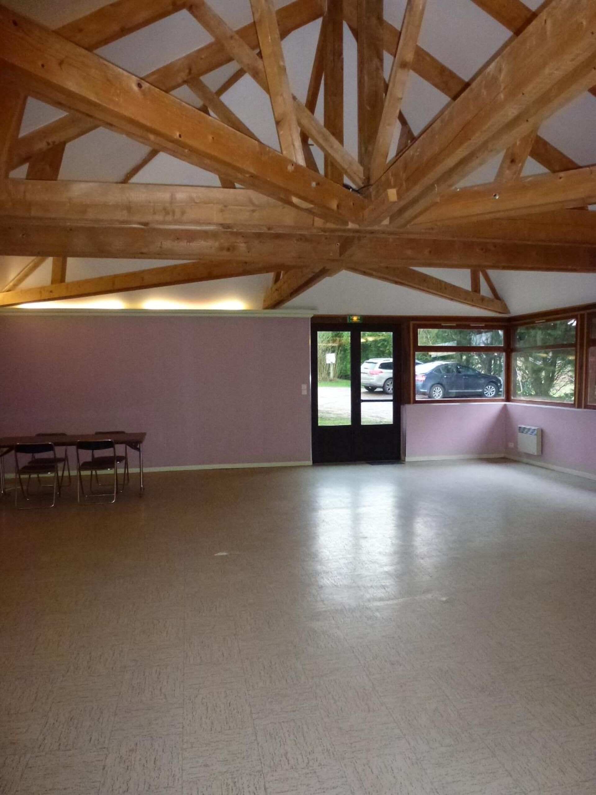 A spacious room with a high wooden ceiling, a door leading outside, and a view of parked cars through large windows.
